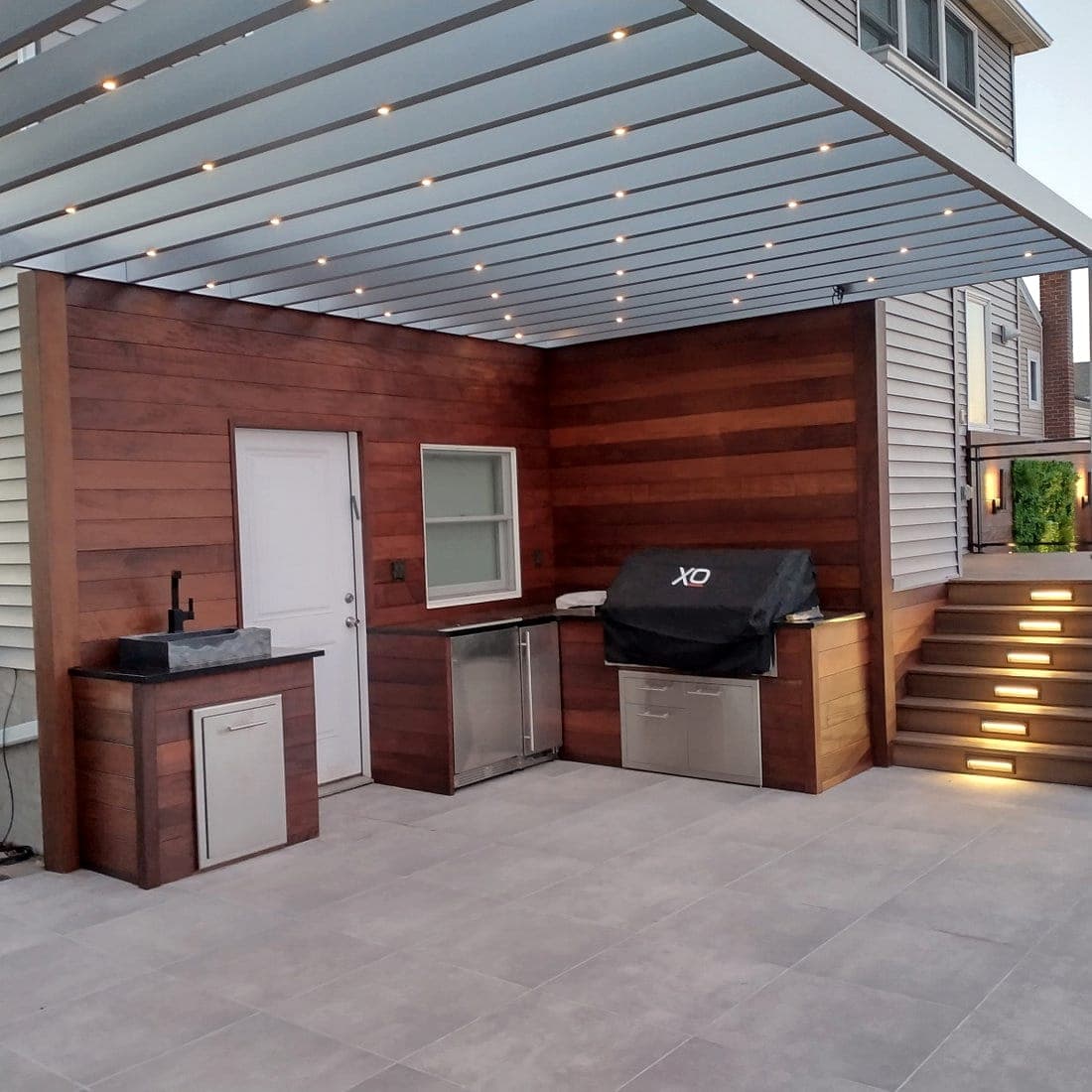 Outdoor kitchen and bar under a louvered pergola, lit at dusk with downlights and IPE wood walls